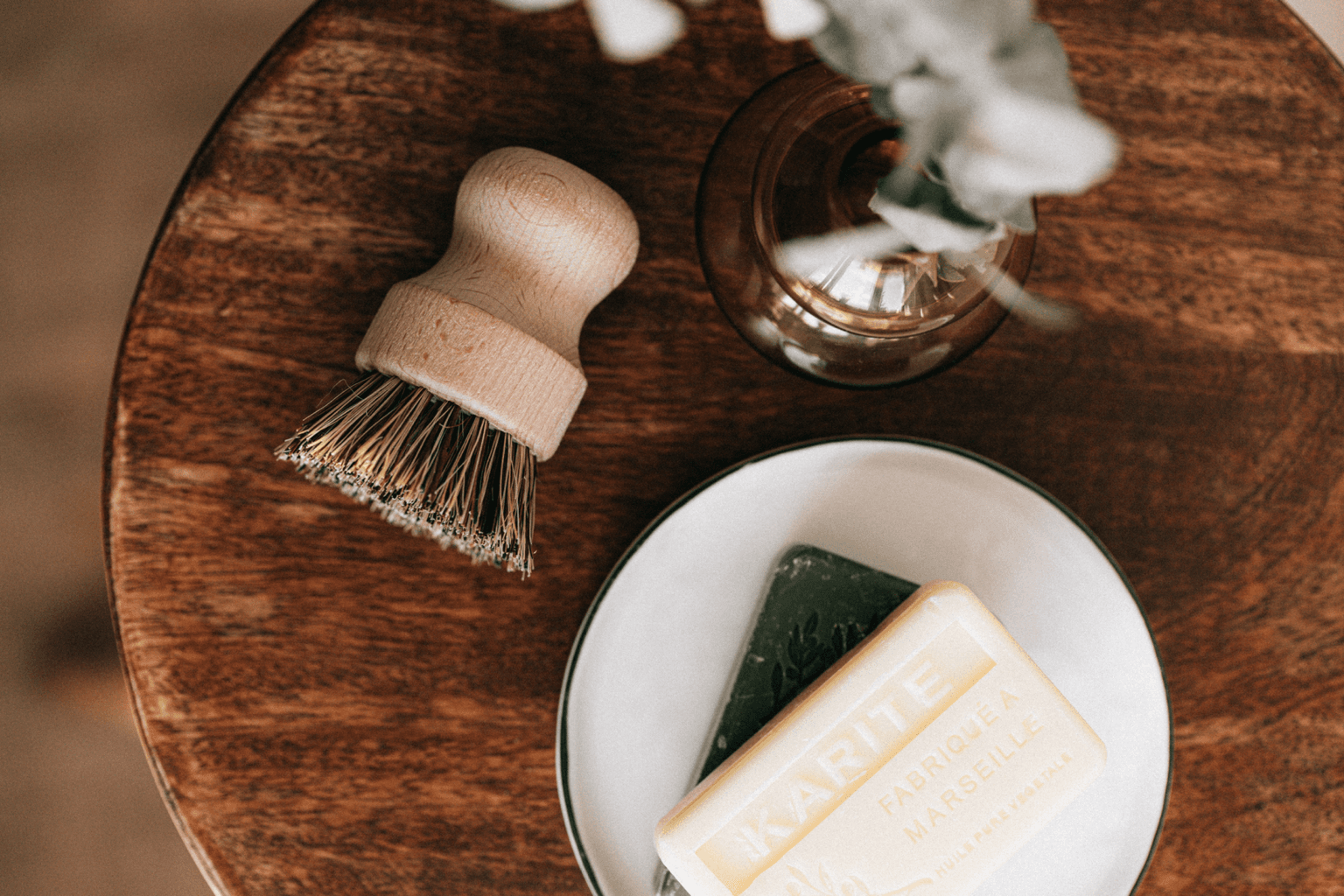 Wooden bristle brush beside a white dish holding a green soap and a cream Karité Fabriqué à Marseille bar on a dark wood table.