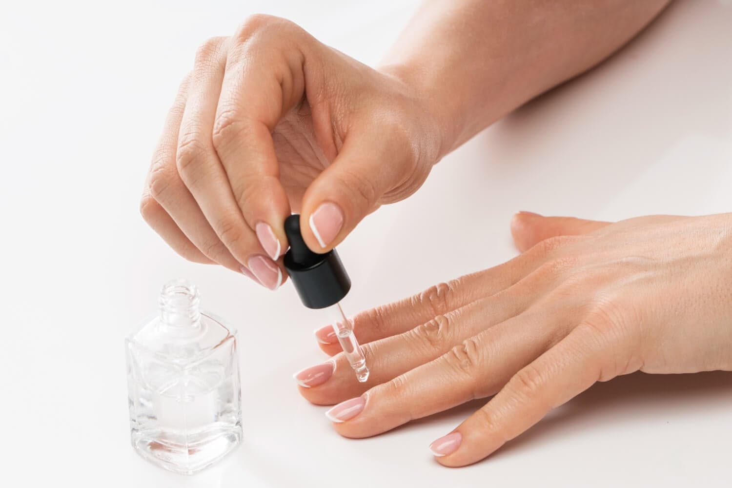 Hands with a French manicure applying clear oil from an unlabelled glass bottle's brush onto a fingernail cuticle.