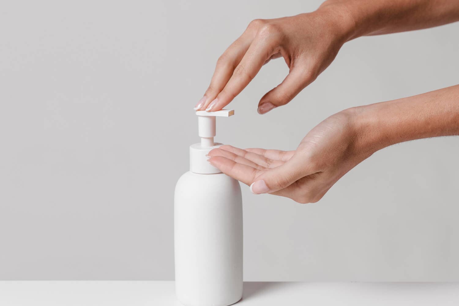 Hands pressing the pump of an unlabelled white soap dispenser bottle on a white surface against a grey background.