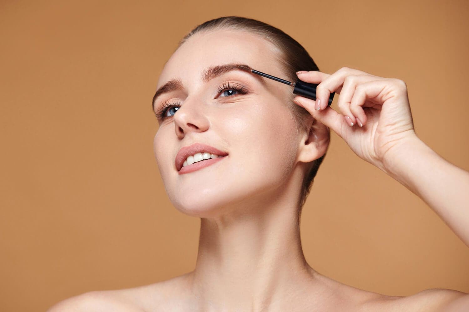 Smiling woman with slicked-back hair brushing her eyebrow with an unbranded black spoolie wand against a warm beige backdrop.