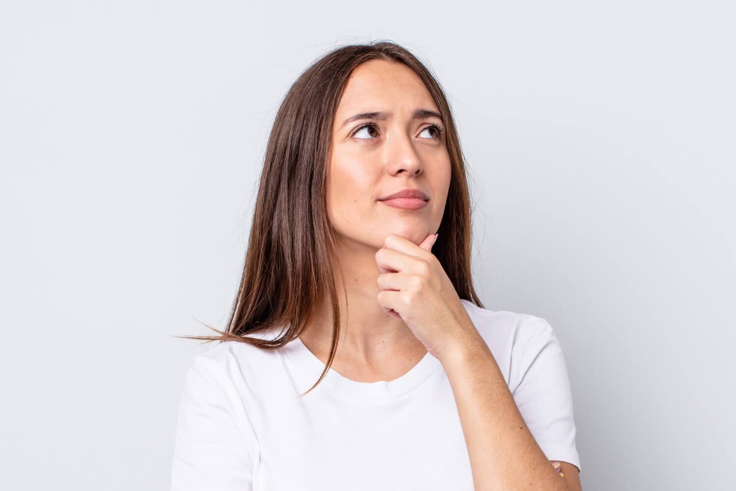 Woman in a white T-shirt with long brown hair, hand on her chin, looking up doubtfully against a plain grey background.