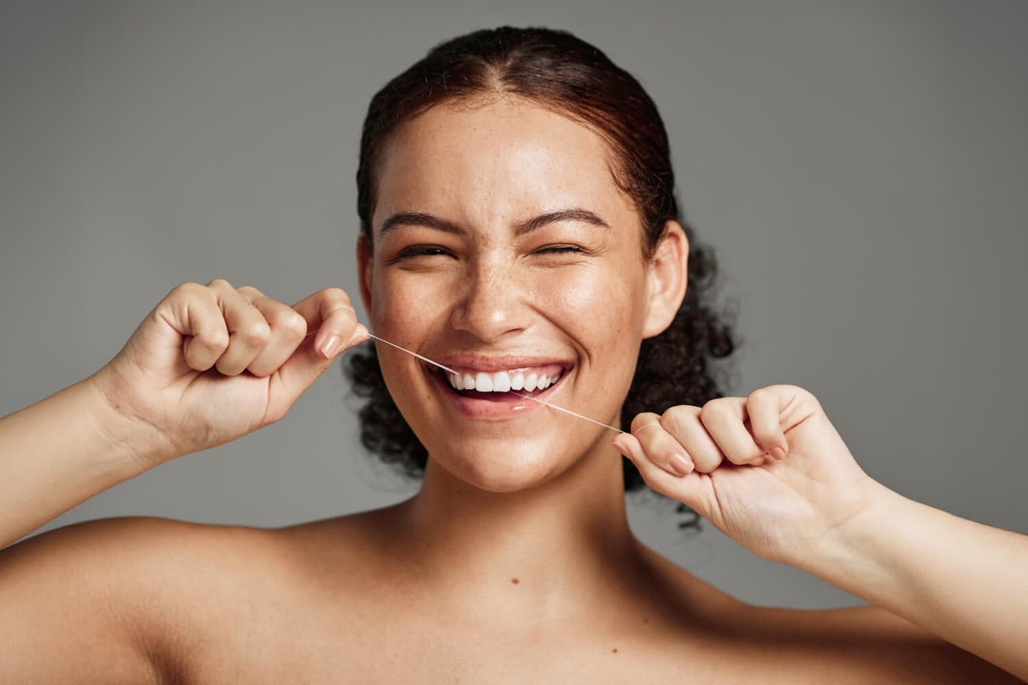 Smiling woman with freckles and dark curly hair holding a strand of dental floss taut against her teeth, on a grey background.